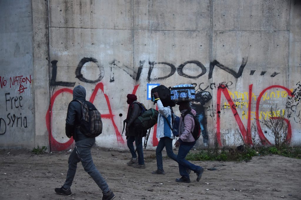 2016-10-24 06:38:07 TOPSHOT - Migrants with luggage walk past a graffiti on a wall reading "London calling", as they leave the "Jungle" migrant camp, as part of an evacuation operation planned to clear the camp in Calais, northern France, on October 24, 2016. French authorities began on October 24, 2016 moving thousands of people out of the notorious Calais Jungle before demolishing the camp that has served as a launchpad for attempts to sneak into Britain. Migrants lugging meagre belongings boarded buses taking them away from Calais' "Jungle" under a French plan to raze the notorious camp and symbol of Europe's refugee crisis. / AFP PHOTO / PHILIPPE HUGUEN