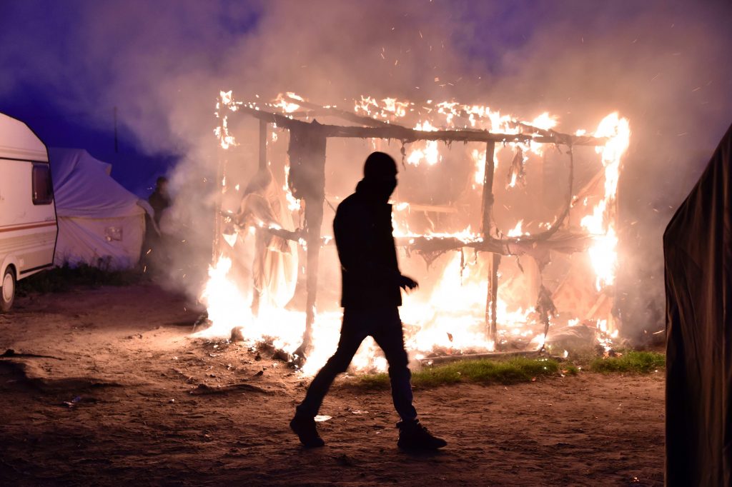 2016-10-25 17:13:03 TOPSHOT - A migrant walks past a shack set on fire during the demolition of the Calais "Jungle" camp, in Calais, northern France, on October 25, 2016 as hundreds of migrants boarded buses on the second day of a massive operation to clear the squalid settlement. More than 1,900 left the slum on October 24, ahead of work to tear down the makeshift shelters and eateries in the camp that has become a symbol of Europe's refugee crisis. / AFP PHOTO / PHILIPPE HUGUEN