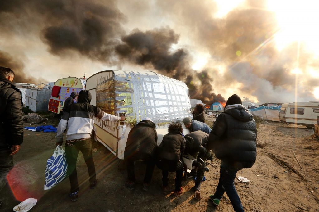 2016-10-26 10:50:48 TOPSHOT - People try to move a caravan as smoke rises from fires at the "Jungle" migrant camp in Calais, northern France, on October 26, 2016, during a massive operation to clear the squalid settlement where 6,000-8,000 people have been living in dire conditions. Fresh fires broke out on October 26 in the "Jungle" camp on the second day of operations to tear down the squalid settlement in northern France, sending migrants fleeing with their meagre belongings. Huge clouds of black smoke billowed over the sprawling camp near Calais where up to 4,000 migrants remain as authorities continue to evacuate residents and dismantle their makeshift dwellings. / AFP PHOTO / François NASCIMBENI